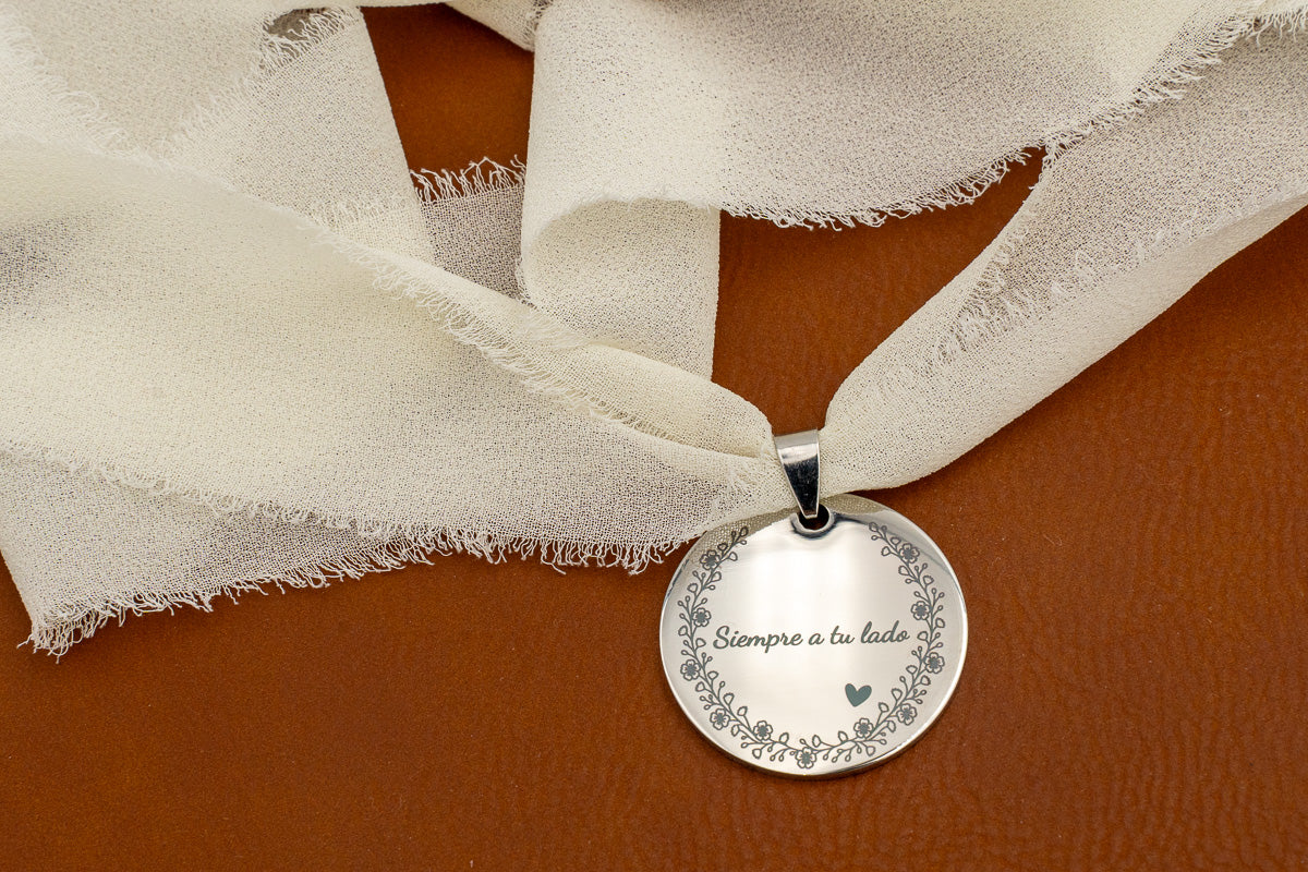 Médaille en acier poli miroir avec la phrase « Toujours à tes côtés » et une bordure florale, nouée avec un ruban en mousseline ivoire