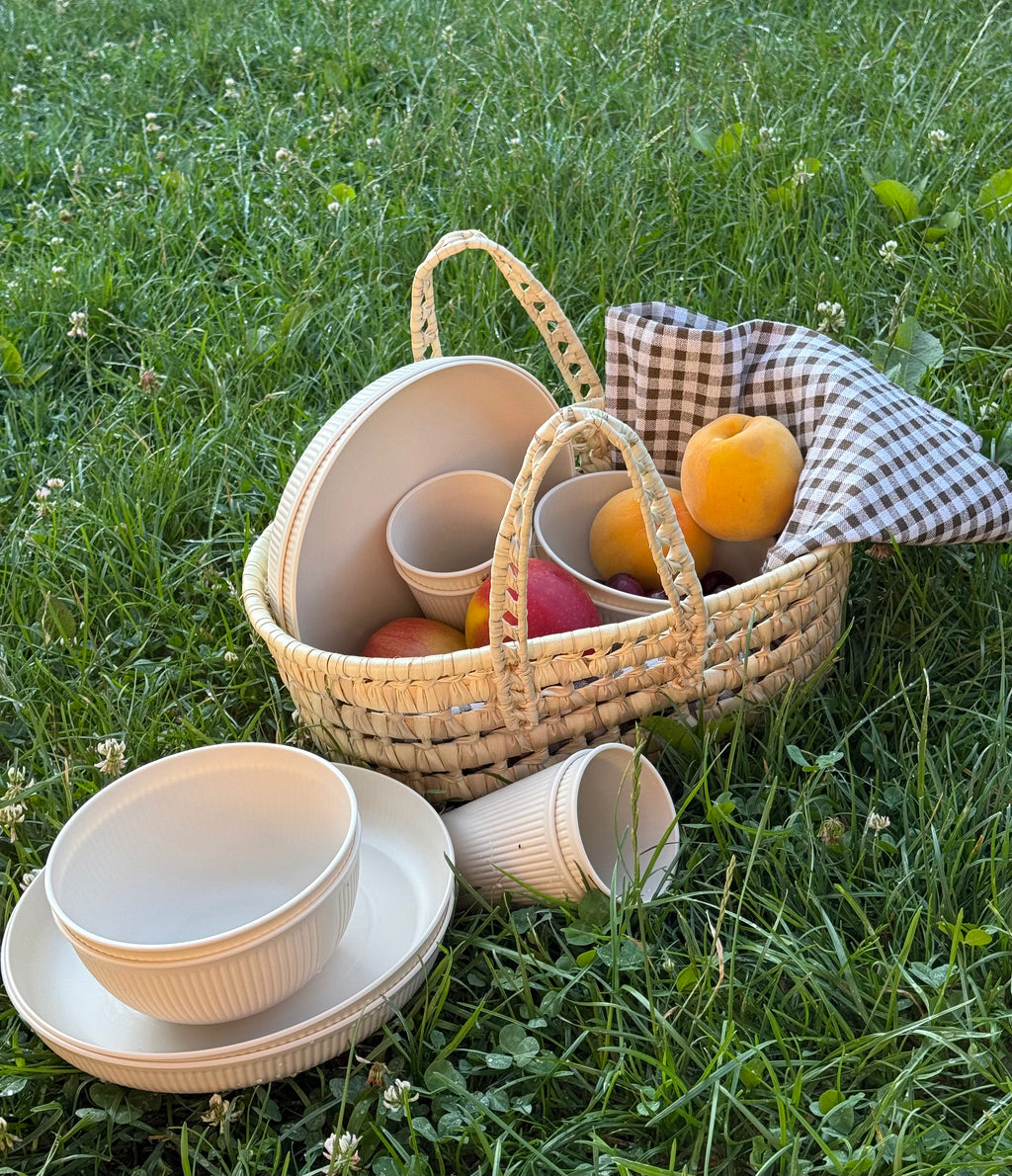 Cesta de rafia con vajilla de polipropileno color crema y fruta fresca sobre césped.