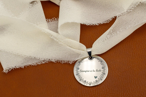Médaille en acier poli miroir avec la phrase « Toujours à tes côtés » et une bordure florale, nouée avec un ruban en mousseline ivoire